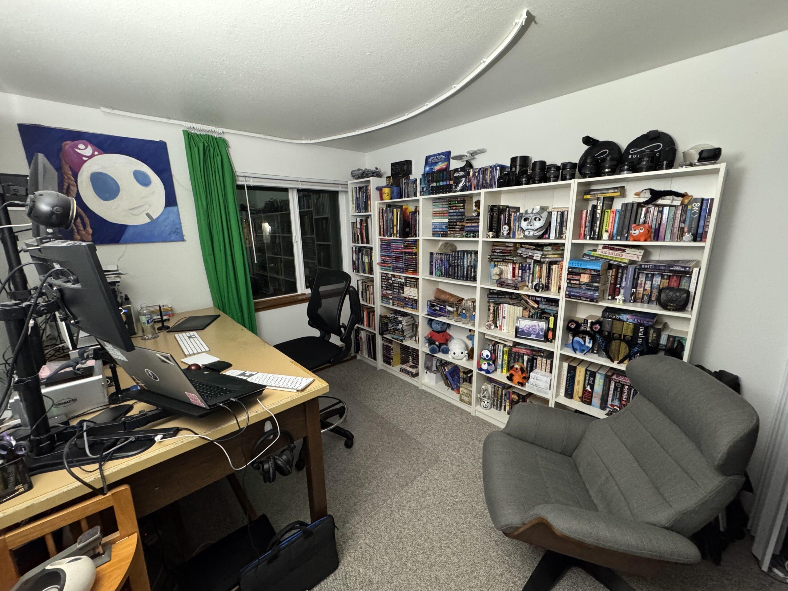 Another view of the same room, with the desk to the left of the image. On the wall opposite the desk is a series of bookcases filled with books and various trinkets and memorabilia. A grey lounge chair sits to the right in the corner of the room. A green curtain hangs to one side from a curve plastic rail mounted on the ceiling.