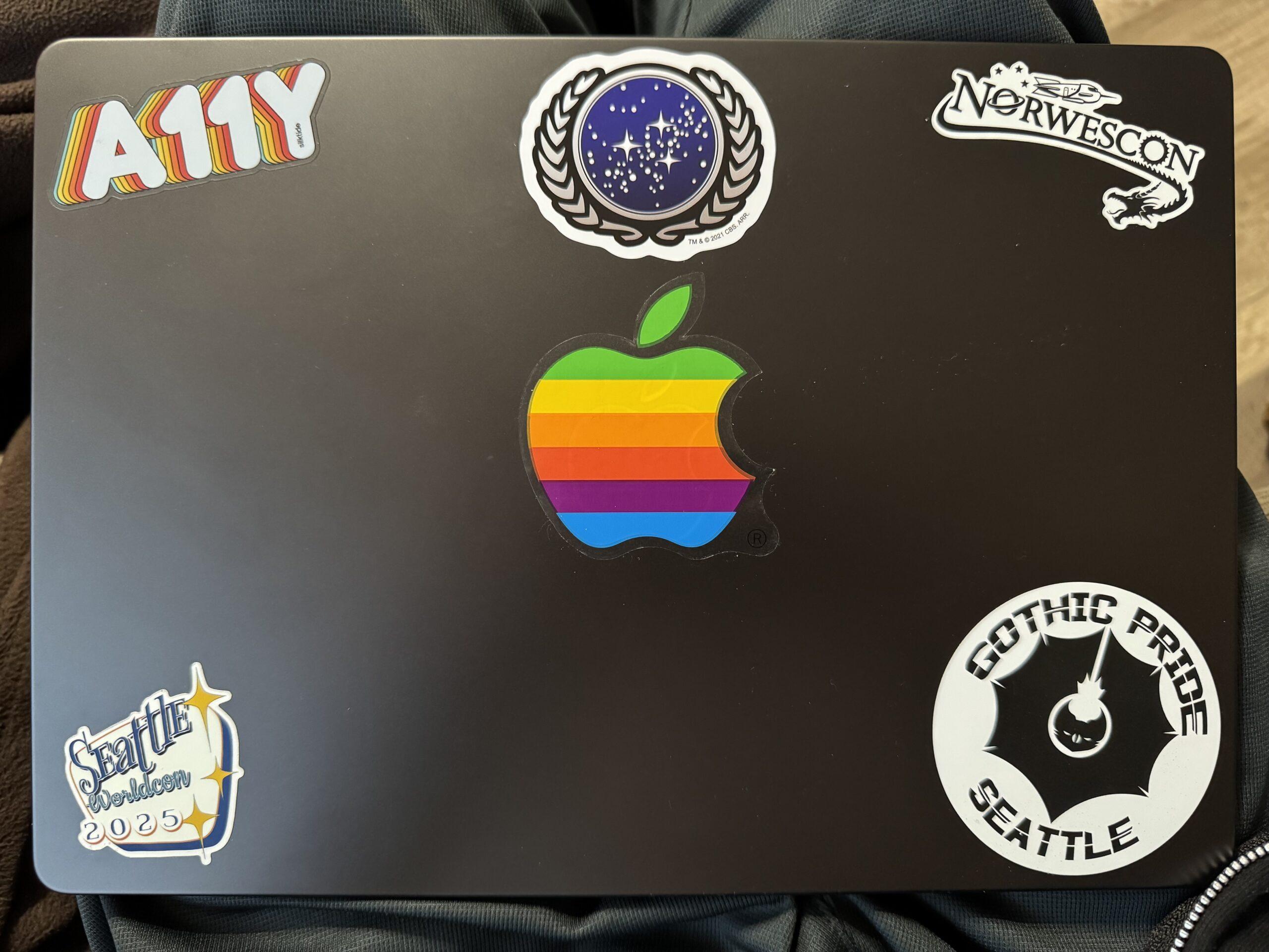 The top of lid of a MacBook Pro with six stickers: A rainbow A11Y, the United Federation of Planets seal, Norwescon, a classic ranbow Apple logo, Seattle Worldcon 2025, and Gothic Pride Seattle.