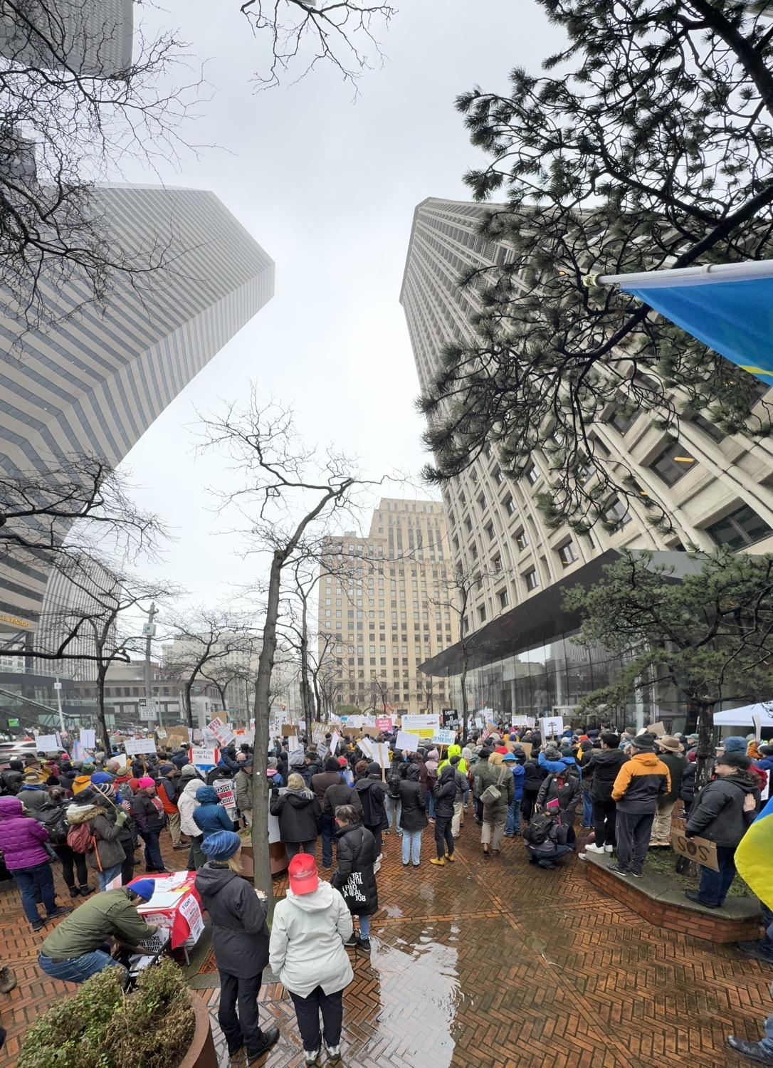 Protesters carrying signs stand on a wet plaza under buildings that seem to loom and bend over them.