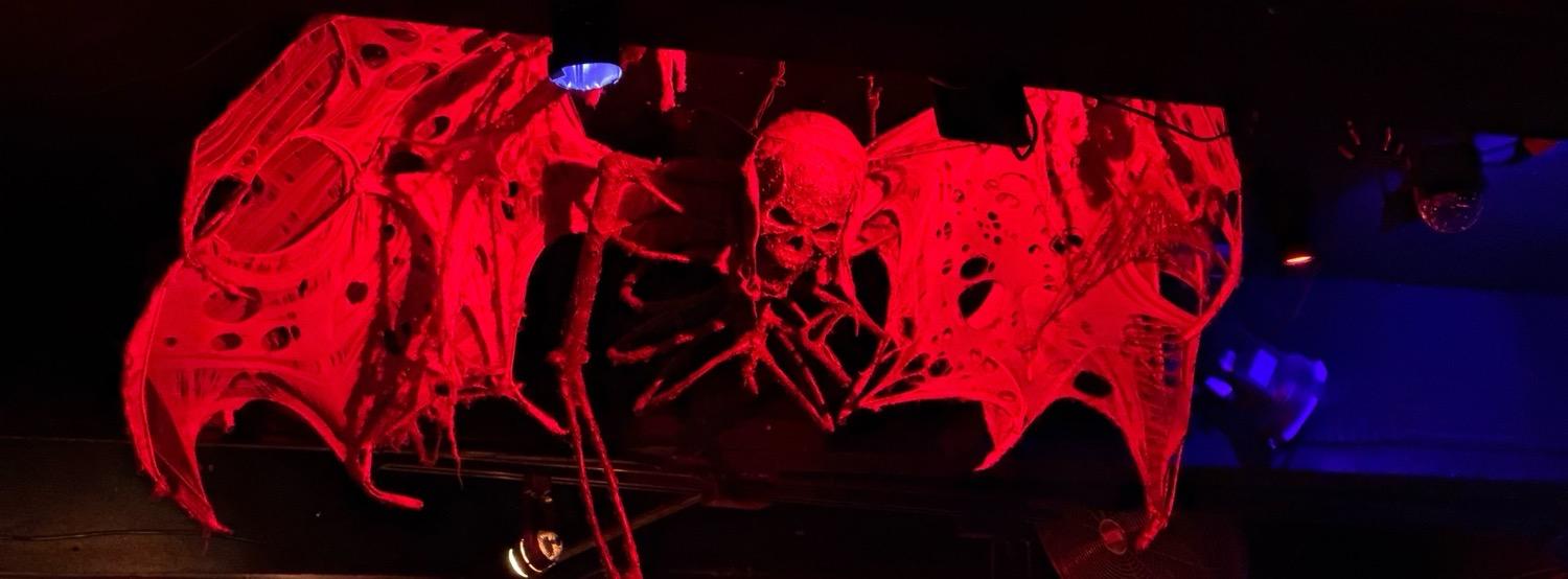 A skeletal ribcage, head, and arms, with tattered bat-like wings, lit all in red, against blue- and amber-toned bits of ceiling in the background.