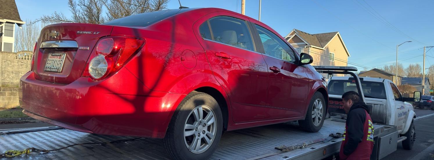 A red Chevy Sonic being strapped onto a flatbed tow truck.