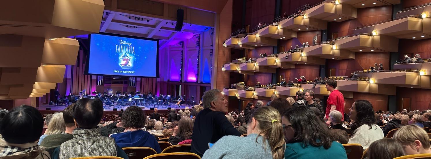 A concert hall filled with people; on the stage are seats for an orchestra below a large screen showing the logo for Disney's Fantasia.