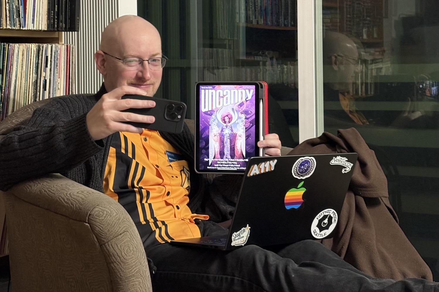 Me sitting in our living room holding my iPhone in my right hand and my iPad with my left hand, and with my MacBook on my lap, as I take a selfie with the latest book I read to blog about it.