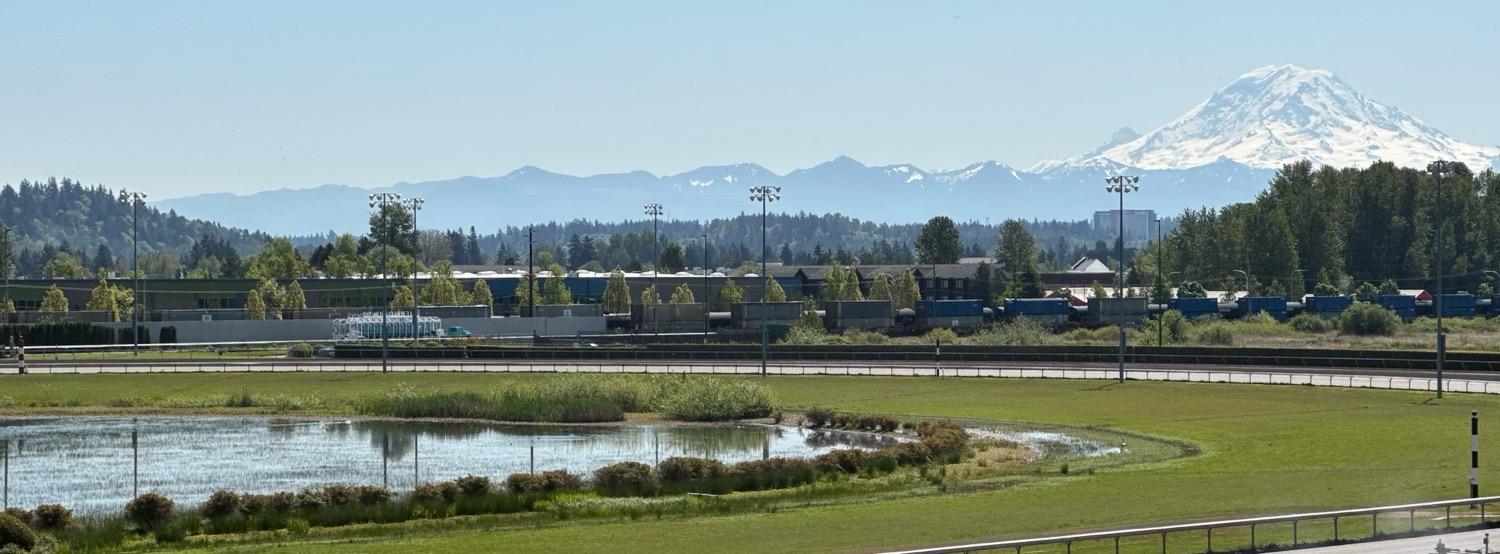 One end of a dirt racetrack circle, with green grass and a pond in the center, with the snow-covered peak of Mt. Rainier rising in the distance in a clear blue sky on a sunny spring day.