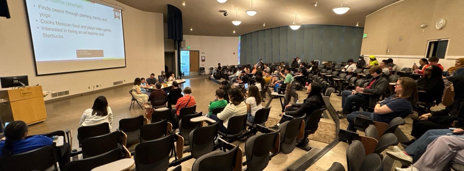 Four students sit at a table at the front of a lecture hall with a presentation screen behind them, in front of an audience of staff, faculty, and fellow students.