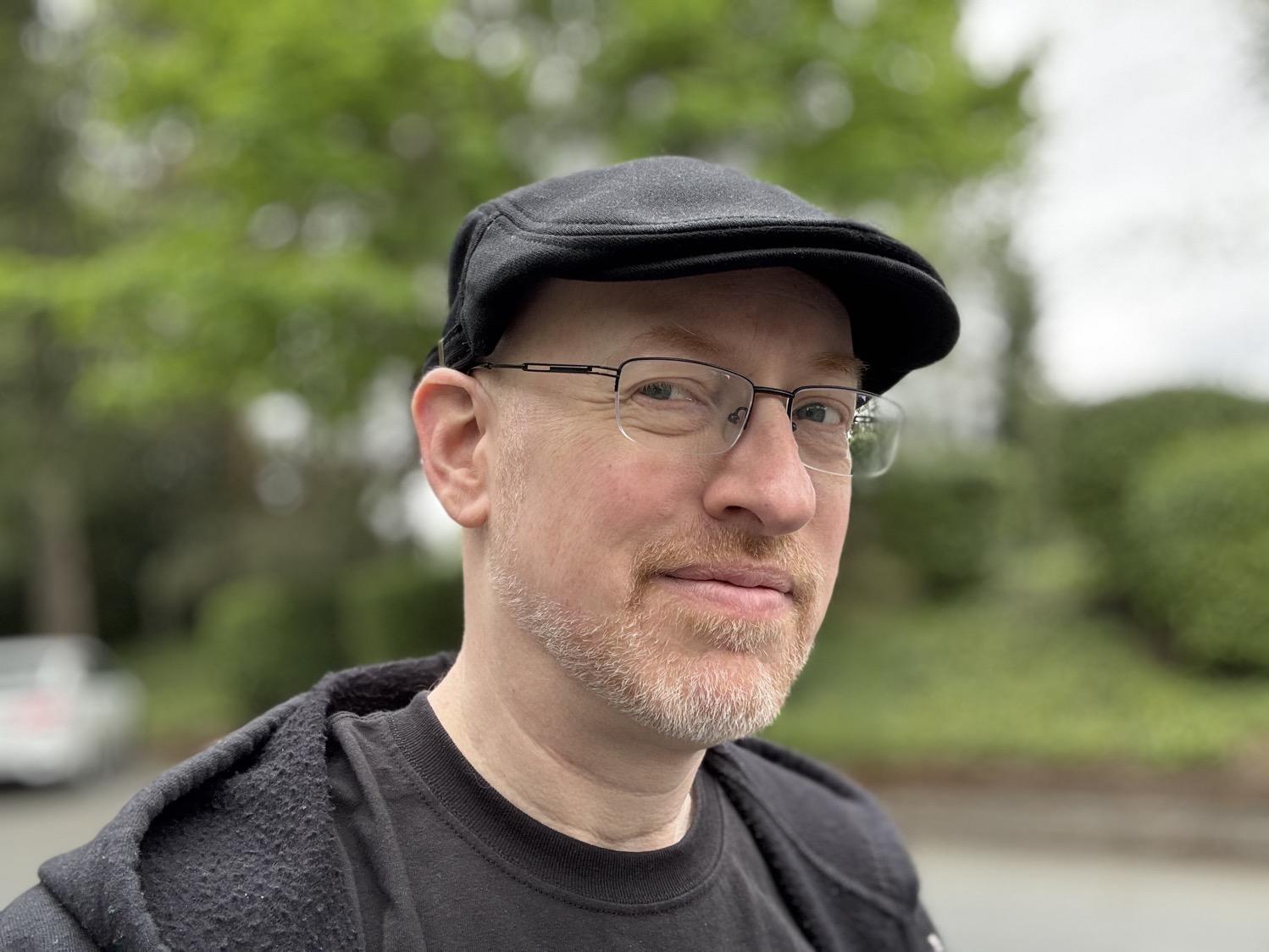 Me, a white man with a short trimmed greying red beard, wearing a black cap, black half-rim glasses, and black shirt and hoodie, standing outside in front of green trees, with a slight smile.