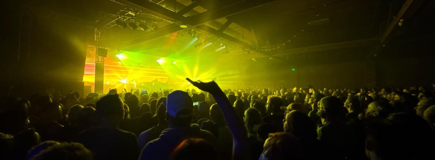 Underworld performing, lit yellows, greens, and oranges, in front of a crowded floor.