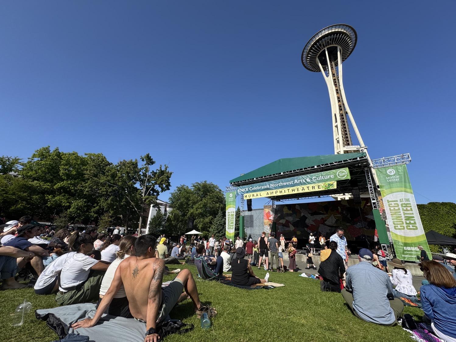 People sit on a lawn in front of an outdoor stage with the Space Needle stretching up into a cloudless blue summer sky.