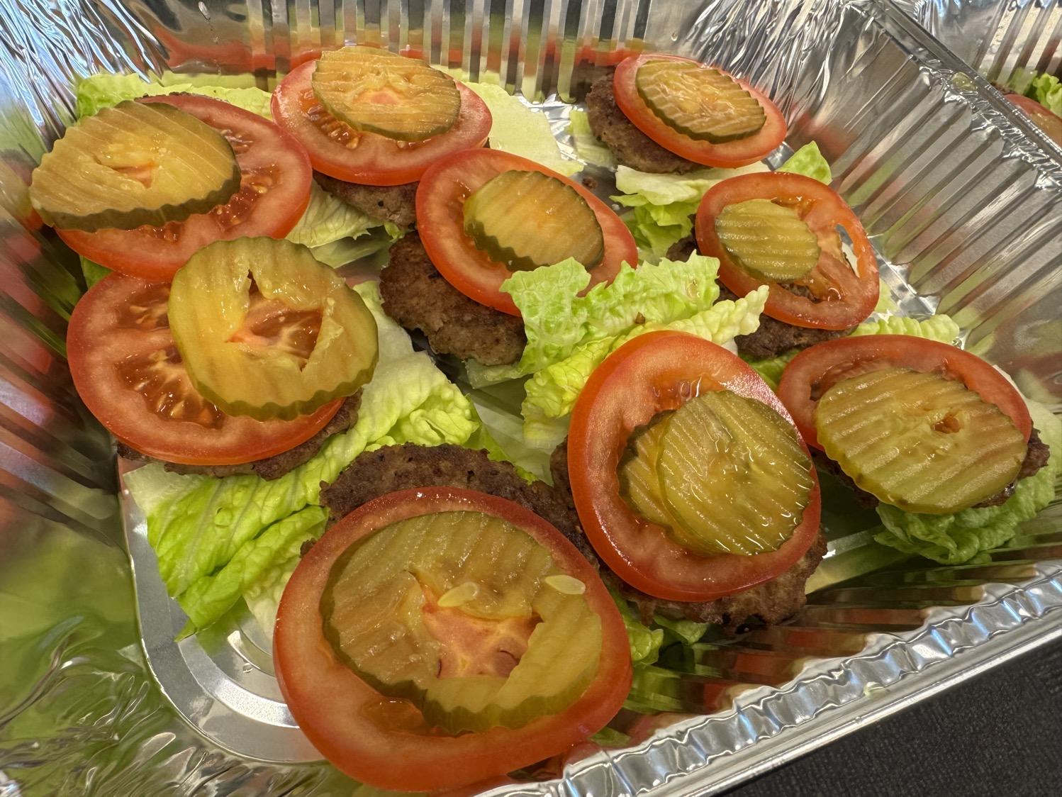 A metal tray with nine snacks made up of stacked lettuce, meat patty, tomato slice, and pickle…and nothing else.