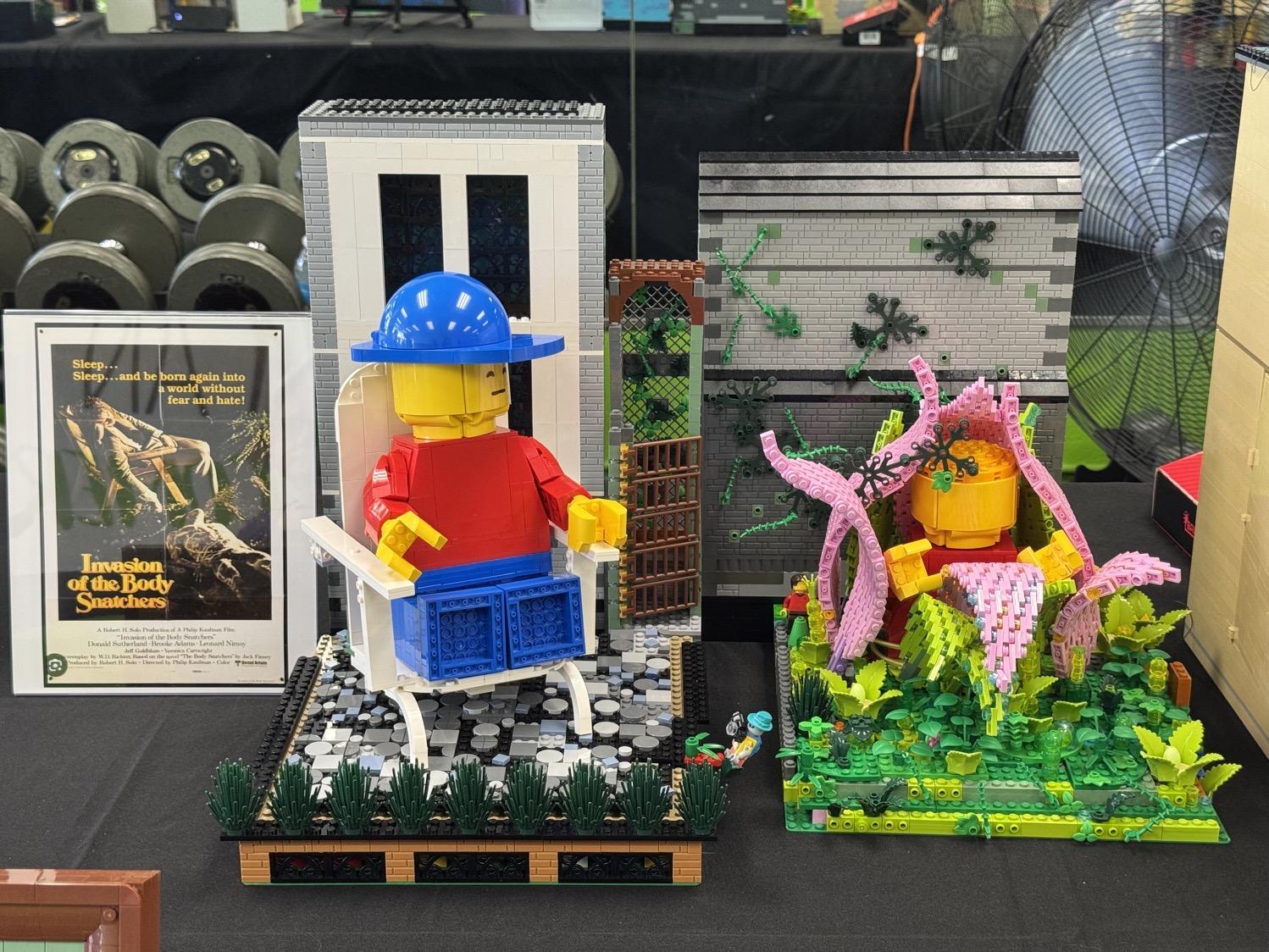 A large size Lego person sitting in a lawn chair next to a plant with another figure emerging from it, both next to a poster for the classic sci-fi horror film Invasion of the Body Snatchers.
