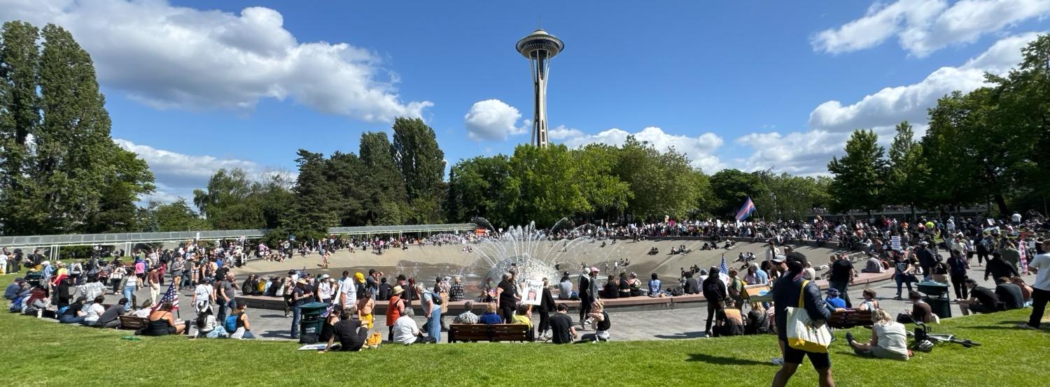 Wide panoramic shot of the Seattle Center's International Fountain crowded with post-march protesters, with the Space Needle in the background under a blue sky with fluffy white clouds.