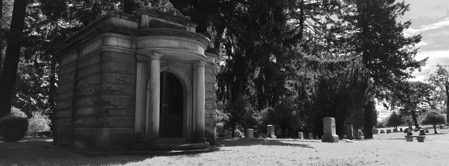 A stone mausoleum next to rows of old gravestones with trees in the background.