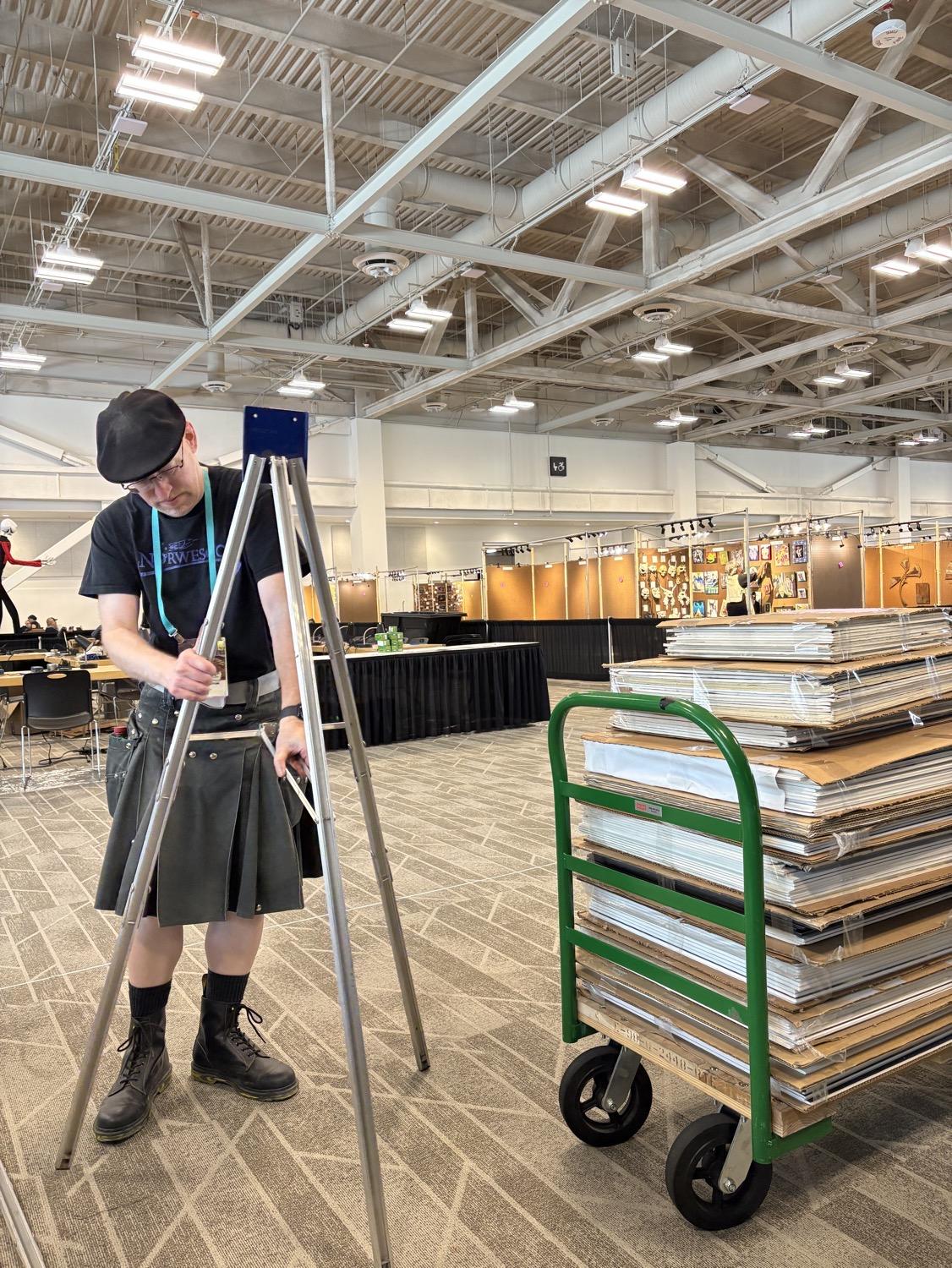 IMG_0710.jpeg Me setting up an aluminum tripod easel in a huge convention center room; next to me is a wheeled pushcart stacked about three feet high with cardboard wrapped bundles.