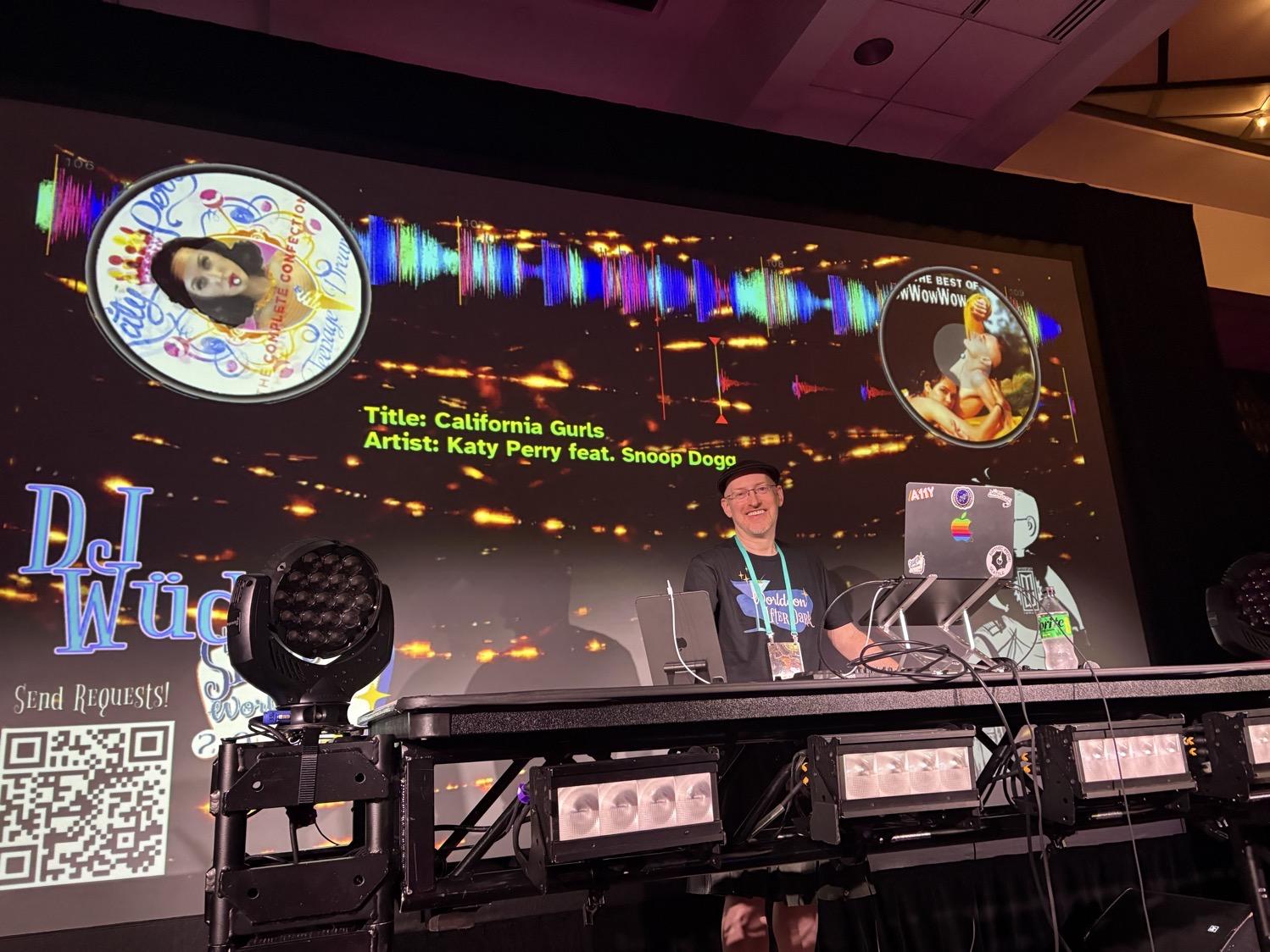 Me standing at a table in front of a MacBook on a stand, in front of a large screen with fancy graphics including cover art and audio waveforms of the songs being played.