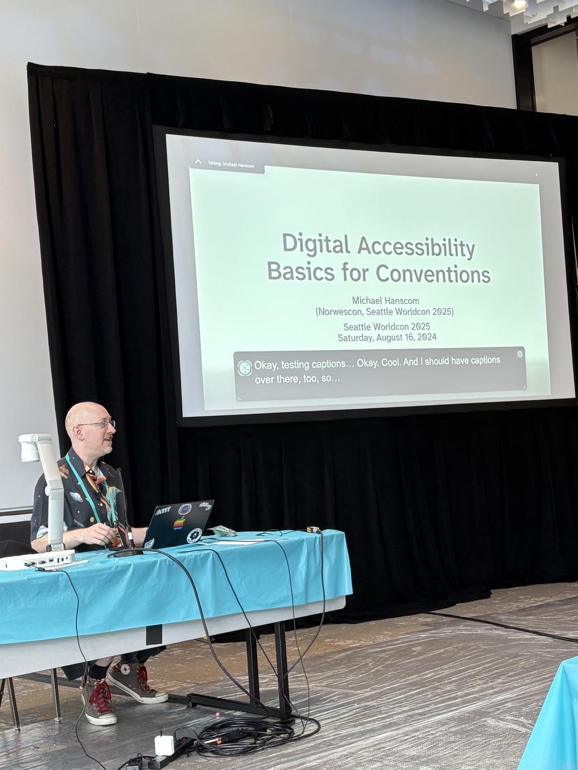 IMG_0960.jpeg Me sitting at a table behind my computer, in front of a large screen displaying a slide with the title 'Digital Accessibility Basics for Conventions' and live captions at the bottom of the screen showing me testing the captioning.
