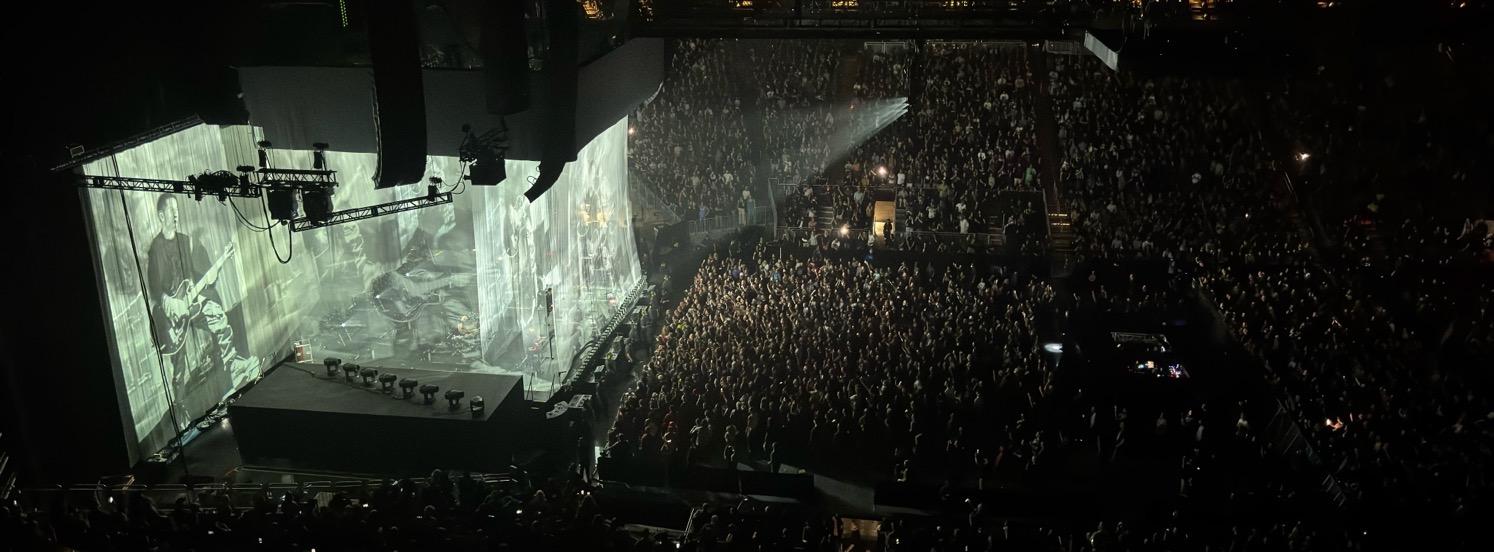 IMG_4656.jpeg The arena filled with people, with the stage at the left of the image, as the band plays behind fabric screens with live images of the band being projected onto them.