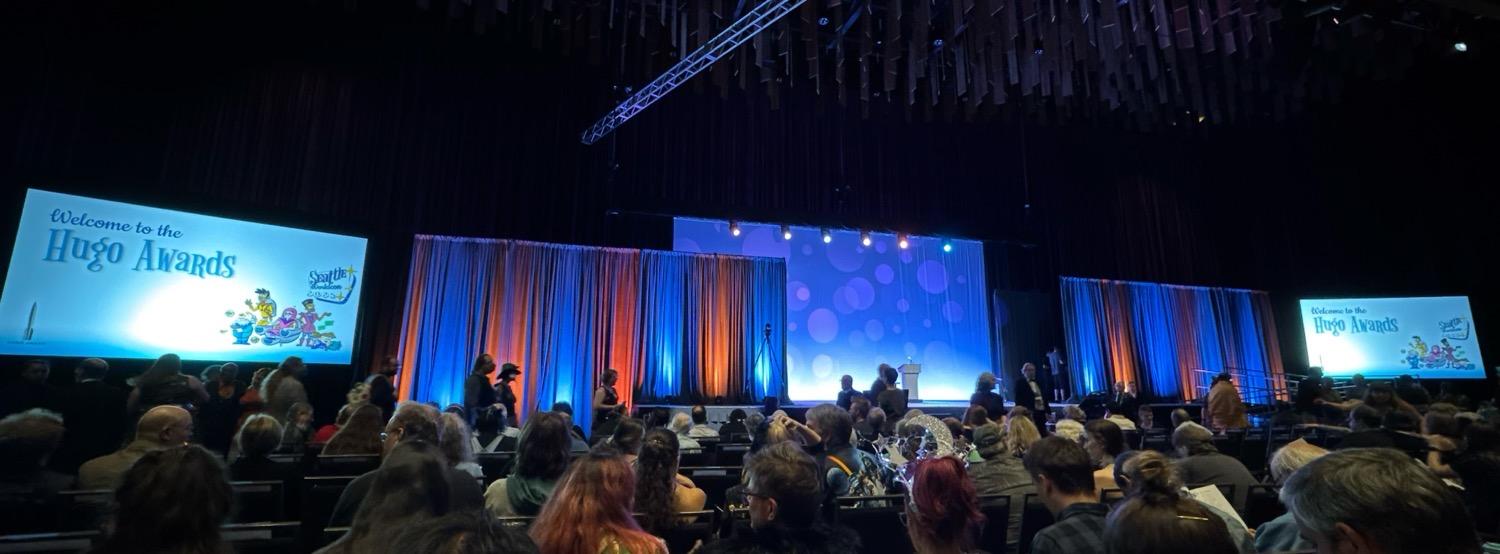 IMG_4714.jpeg Wide shot of the Hugo award ceremony stage, lit in blues and with blue and orange curtains to the sides, with large video screens to the far left and right showing a slide that says Welcome to the Hugo Awards.