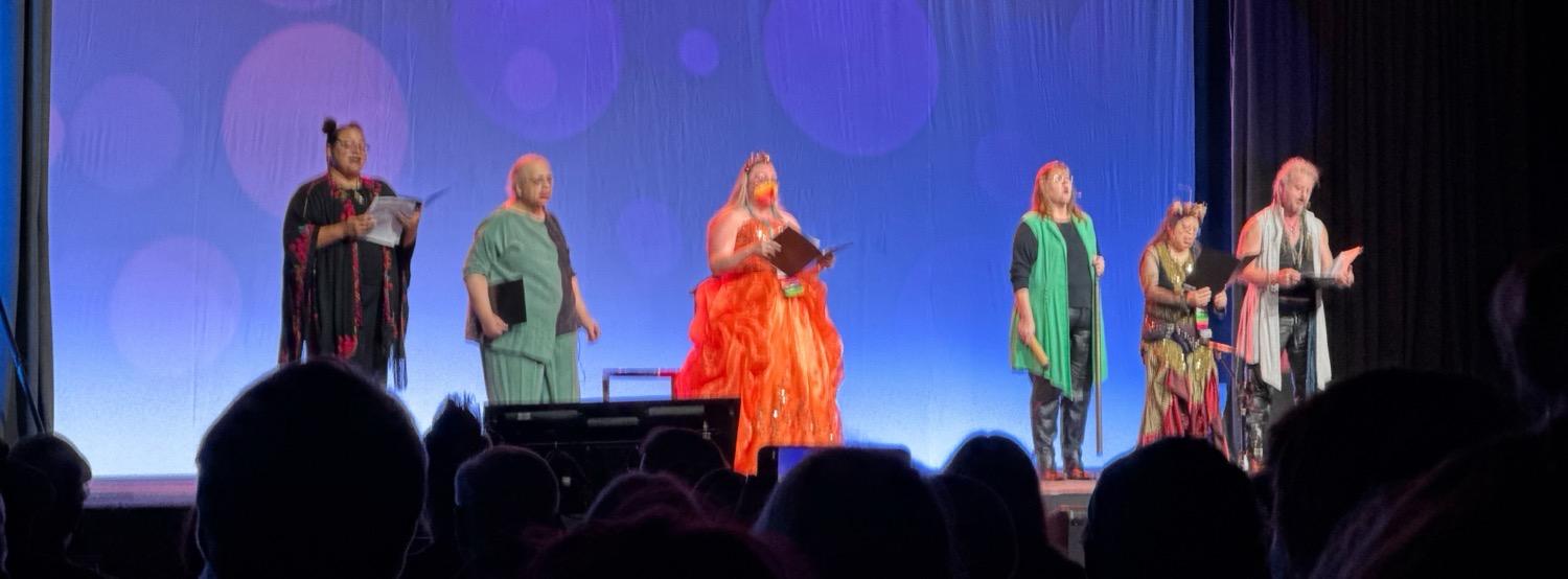 IMG_4717.jpeg Six people in various forms of fancy dress on stage, some holding booklets as they sing.