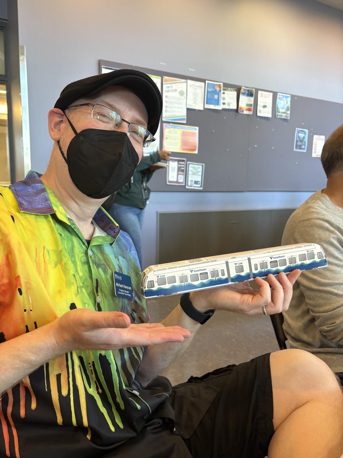 Me holding a paper model of a Seattle Link light rail train car, while wearing a black cap, rainbow shirt, and black face mask.