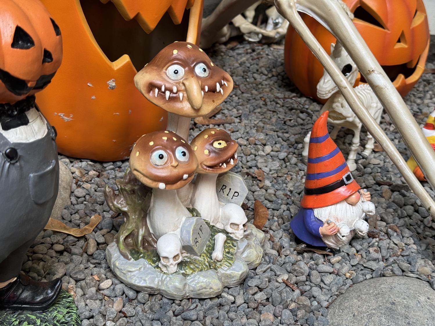 An outdoor decorative figure of three very creepy mushrooms with wide eyes, droopy noses, and wide evil sharp-toothed smiles, with tombstones and bones at their bases.