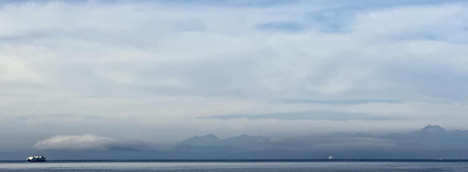 Panoramic shot of the Olympic mountains across Puget Sound, half-shrouded in clouds, under a mostly cloudly sky, with a ferry on the water on the far left of the image.