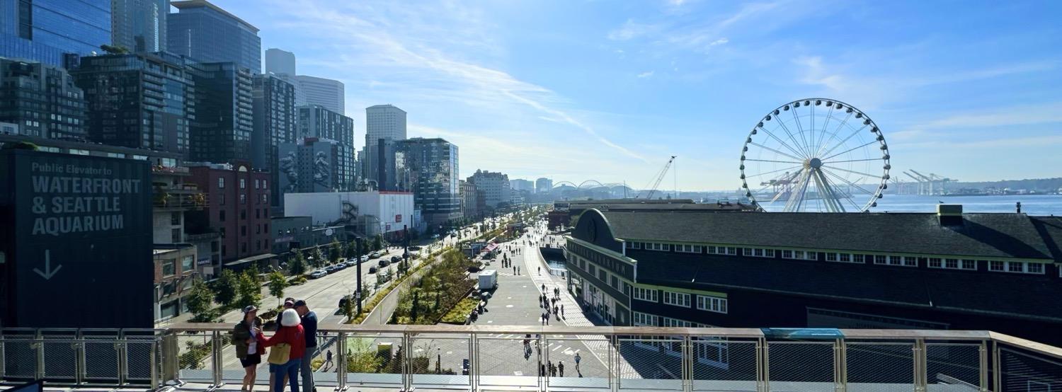 Looking south down the Seattle waterfront from the roof of the new aquarium with the skyline on the left and the Seattle ferris wheel on the right, with people strolling along the sidewalk by the old aquarium building.