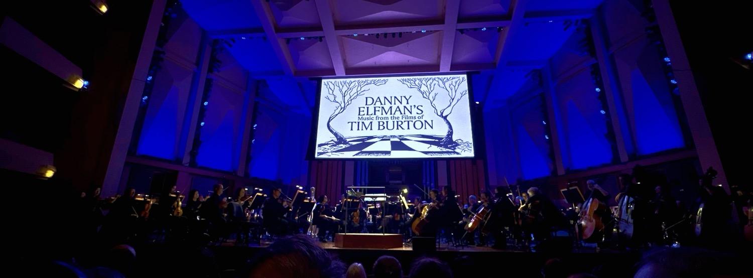 The Seattle Symphony on stage, with blue and purple lights on the walls, and a screen displaying a Tim Burton sketch of two bare trees on a checkerboard landscape and the text, 'Danny Elfman's music from the films of Tim Burton'.