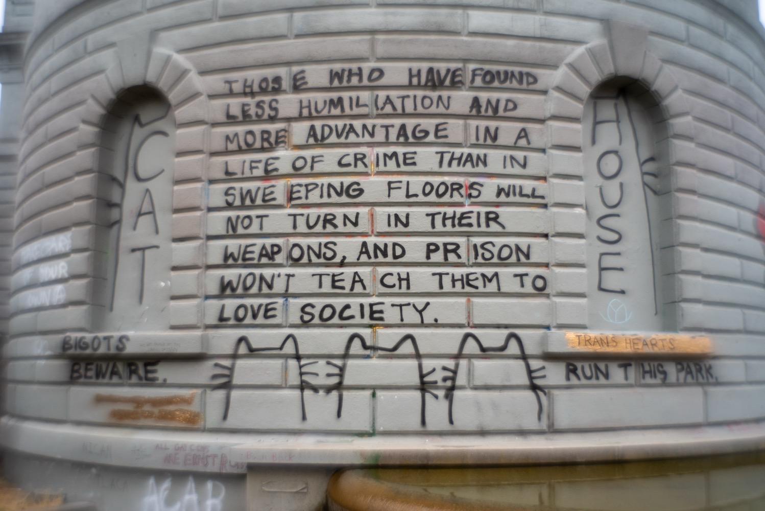 A curved grey brick wall with graffiti that reads, 'those who have found less humiliation and more advantage in a life of crime than in sweeping floors will not turn in their weapons, and prison won't teach them to love society', surrounded by more graffiti of cat heads and text that says, 'bigots beware' and 'trans hearts run this park'.
