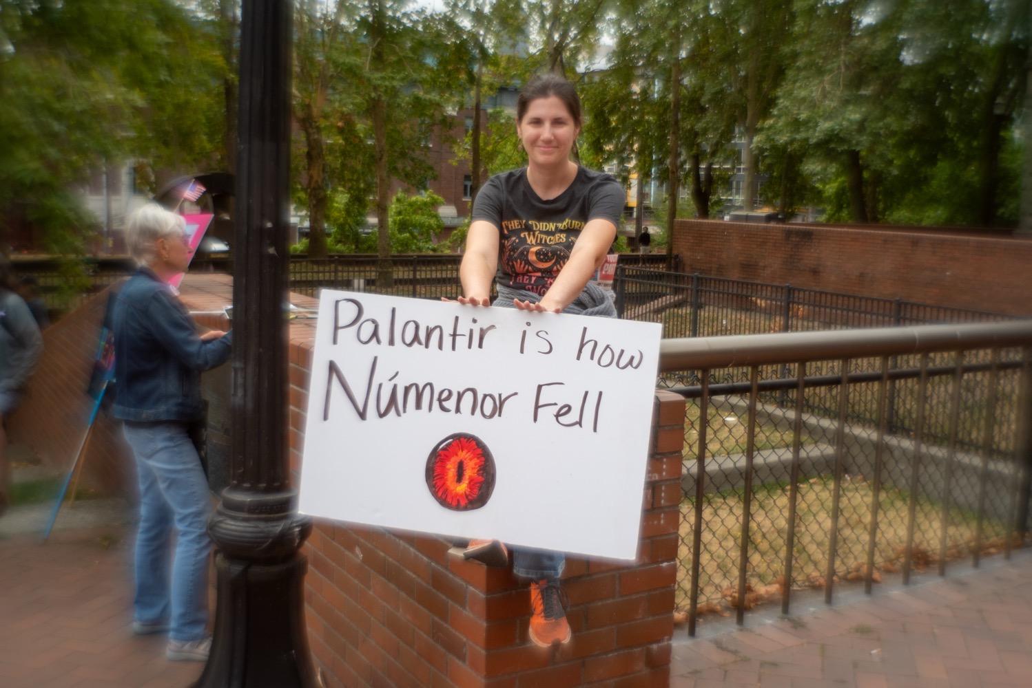 A woman sitting on a brick wall and smiling, holding a sign that says, 'Palantir is how Númenor fell'.
