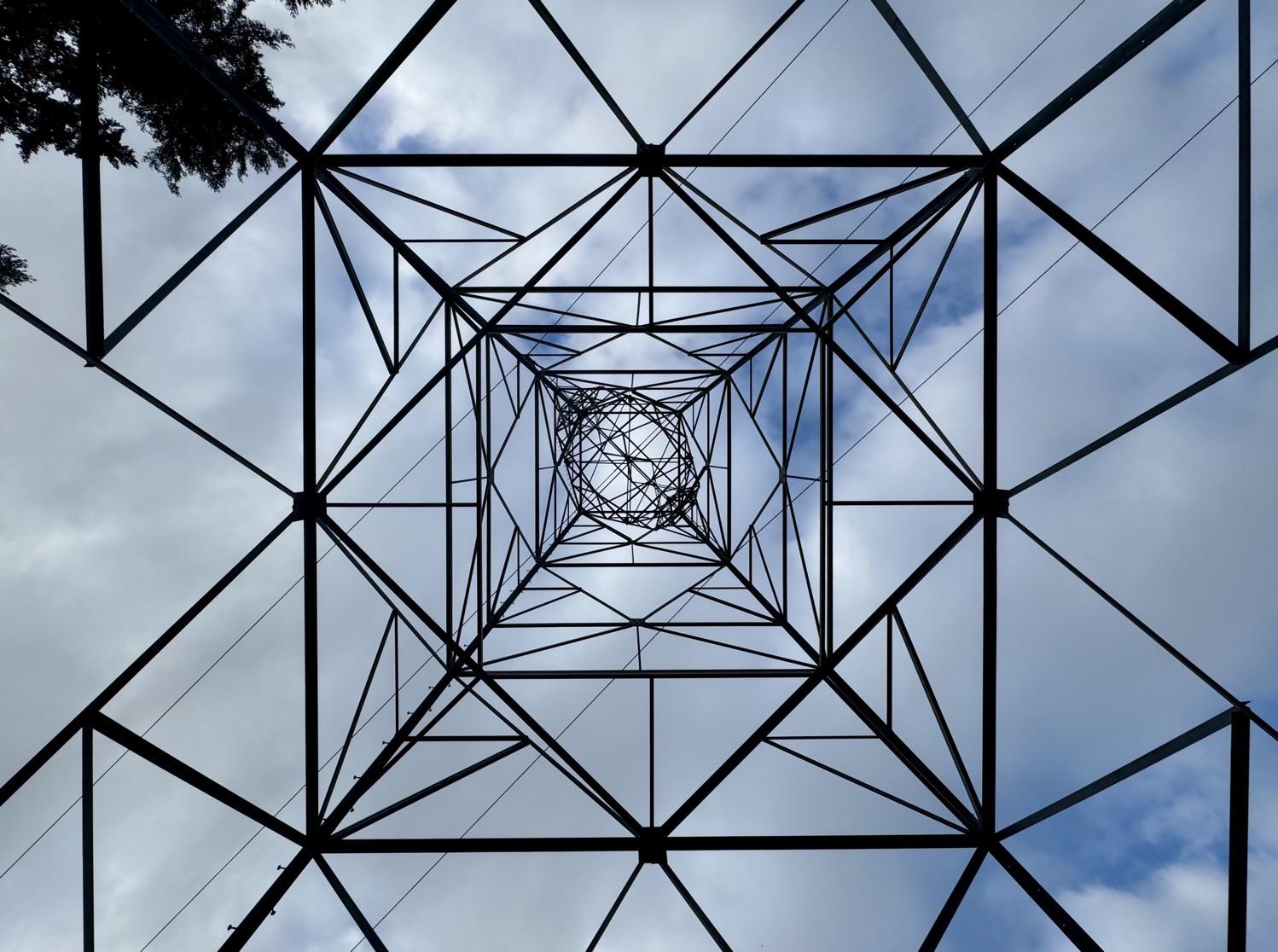 White clouds mostly cover a blue sky above a grid of black horizontal, vertical, and diagonal lines made by the girders of an electrical tower.
