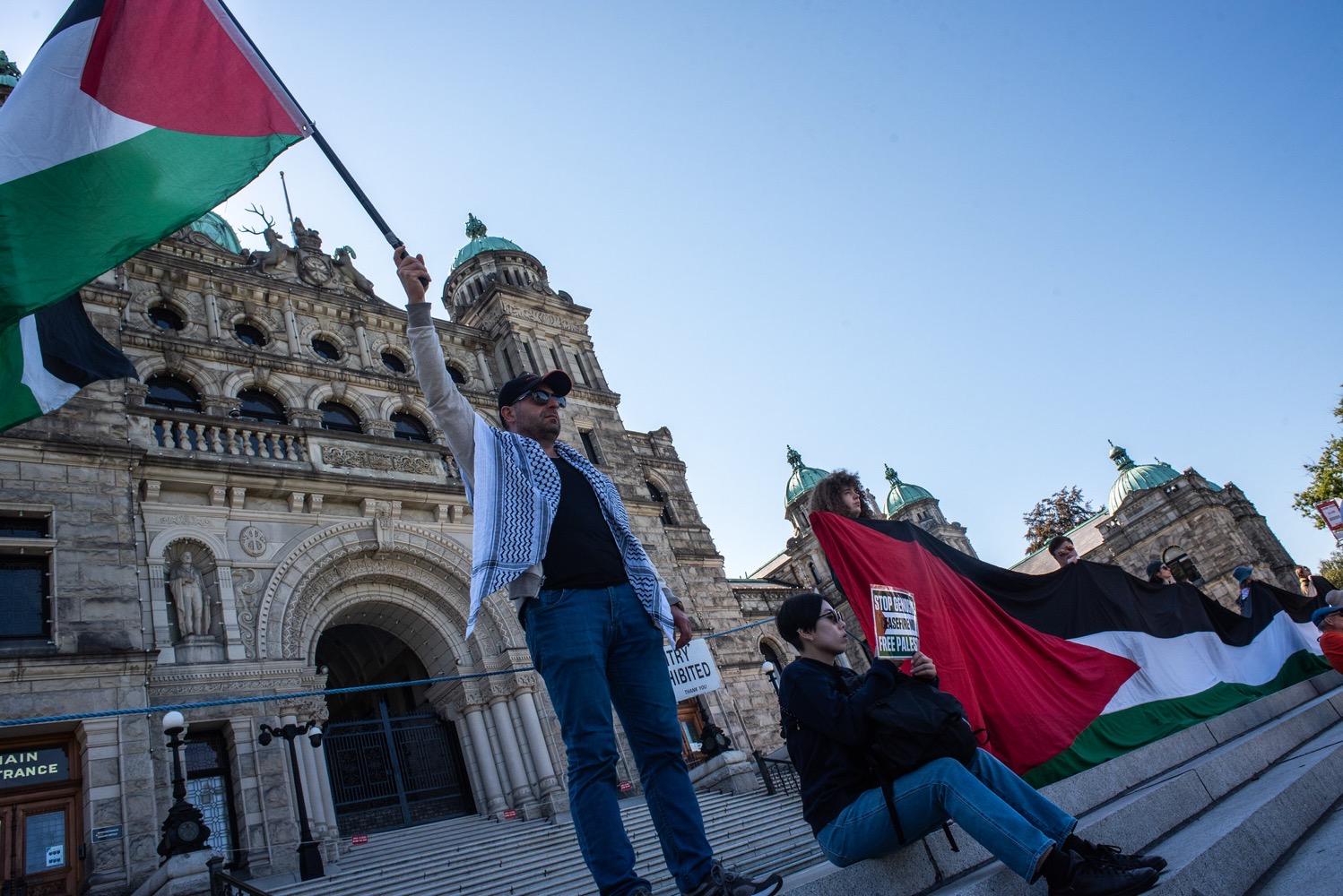 A man wearing a keffiyah and waving a Palestinian flag stands next to a row of people holding an elongated Palestinian flag on the steps in front of Victoria's Parliament building.