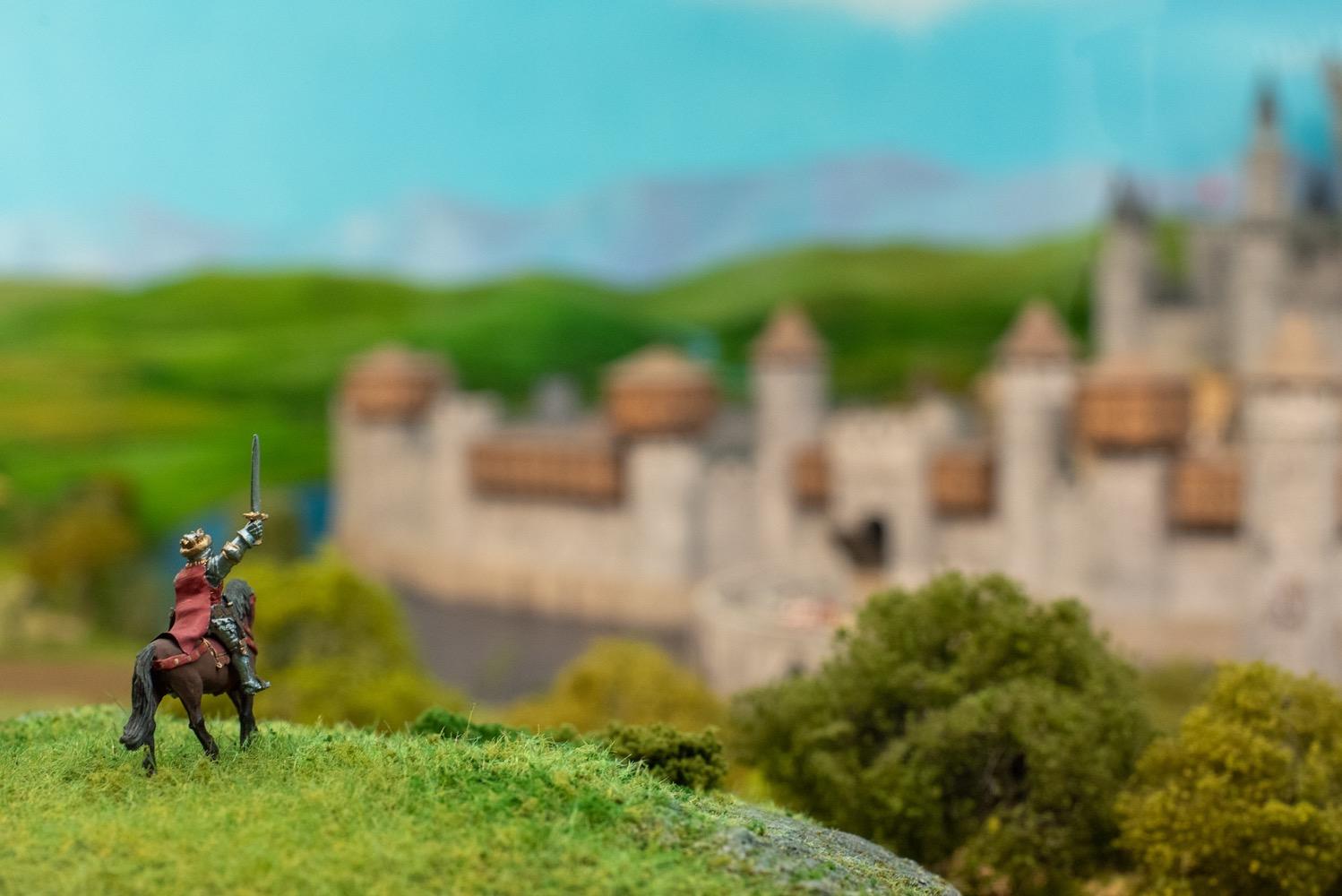 On a hilltop with Camelot visible in the distance, a tiny knight in armor on horseback raises a sword in salute.