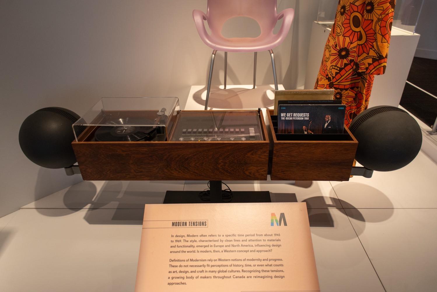 A 1960s stereo on display in a museum. It's made of two wooden cases, one on the right to hold LPs, the other on the left, twice as wide, to hold the turntable and central control panel. Black spherical speakers are on either side. The whole thing is probably about eight feet wide.