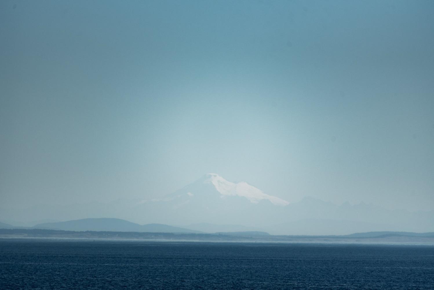 A snow-covered mountain in the distance under a clear blue sky and with flat blue ocean in the foreground.