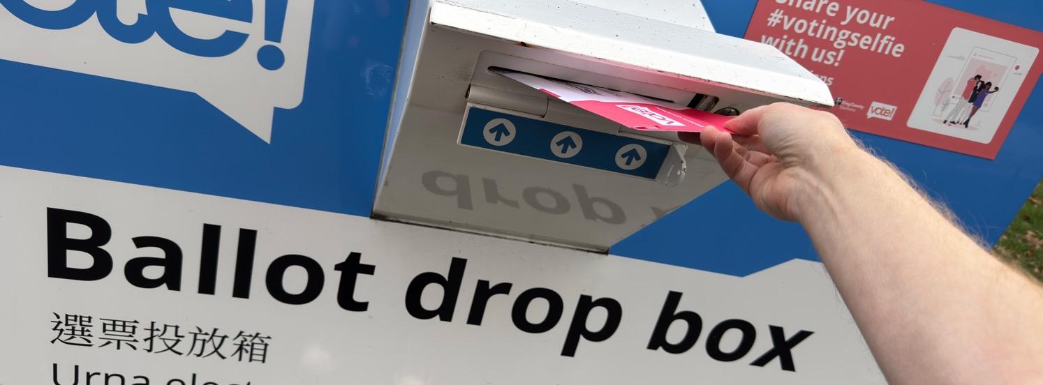 My hand placing a Washington ballot envelope into a blue-and-white King County ballot drop box.