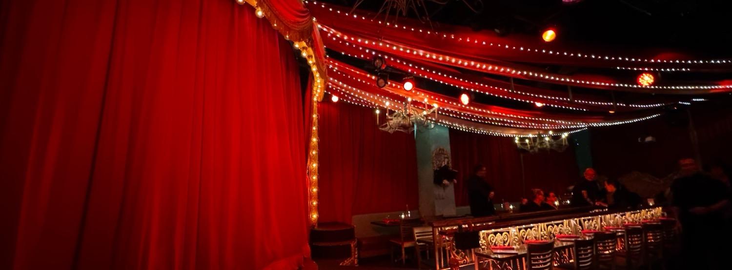 The Can Can room all in red and gold, with a stage on the left with a closed red velvet curtain, red fabric stretched aross the ceiling over strings of lights.