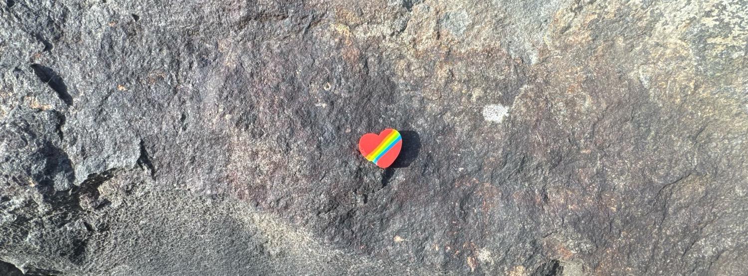 A small red heart with diagonal rainbow stripes against stone.
