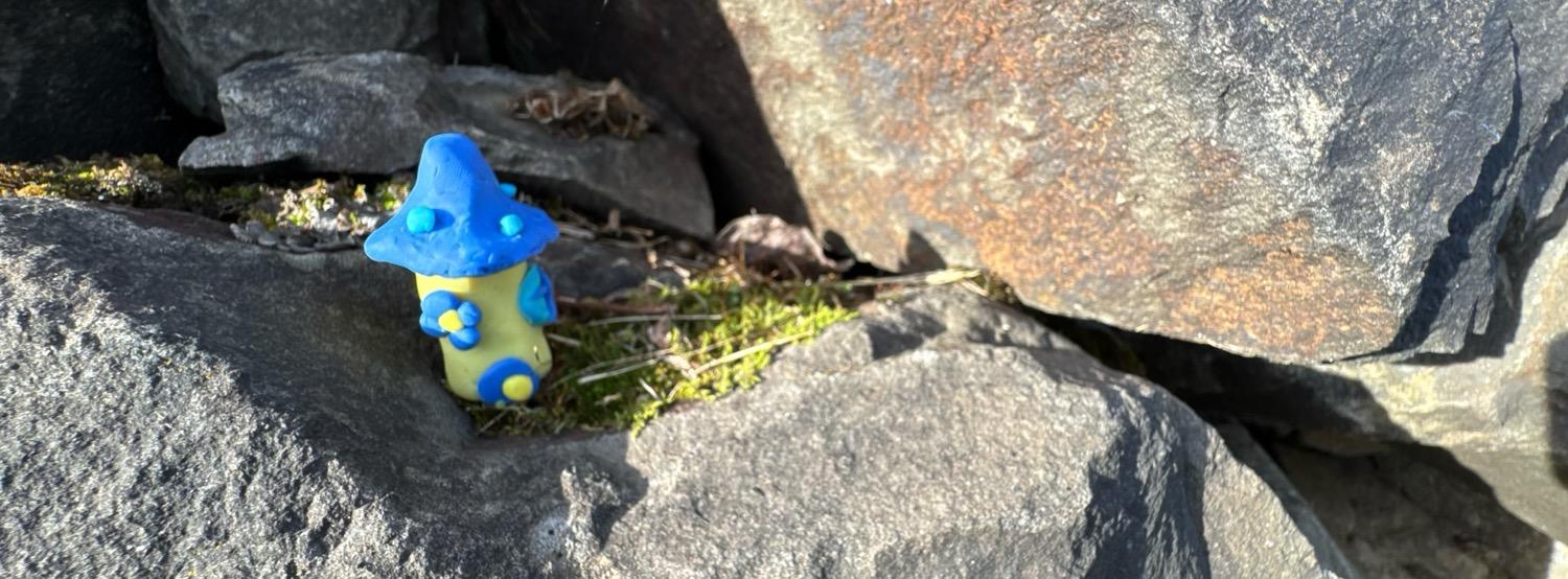 A blue and yellow sculpted clay mushroom house sitting in a crack between stones in a rock wall.