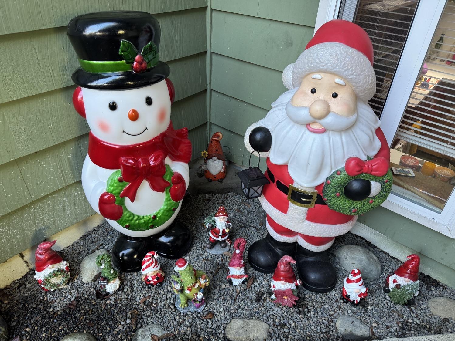 Outdoor plastic snowman and Santa standees in an outdoor graveled corner, with a lineup of holiday-themed gnome figurines at their feet.