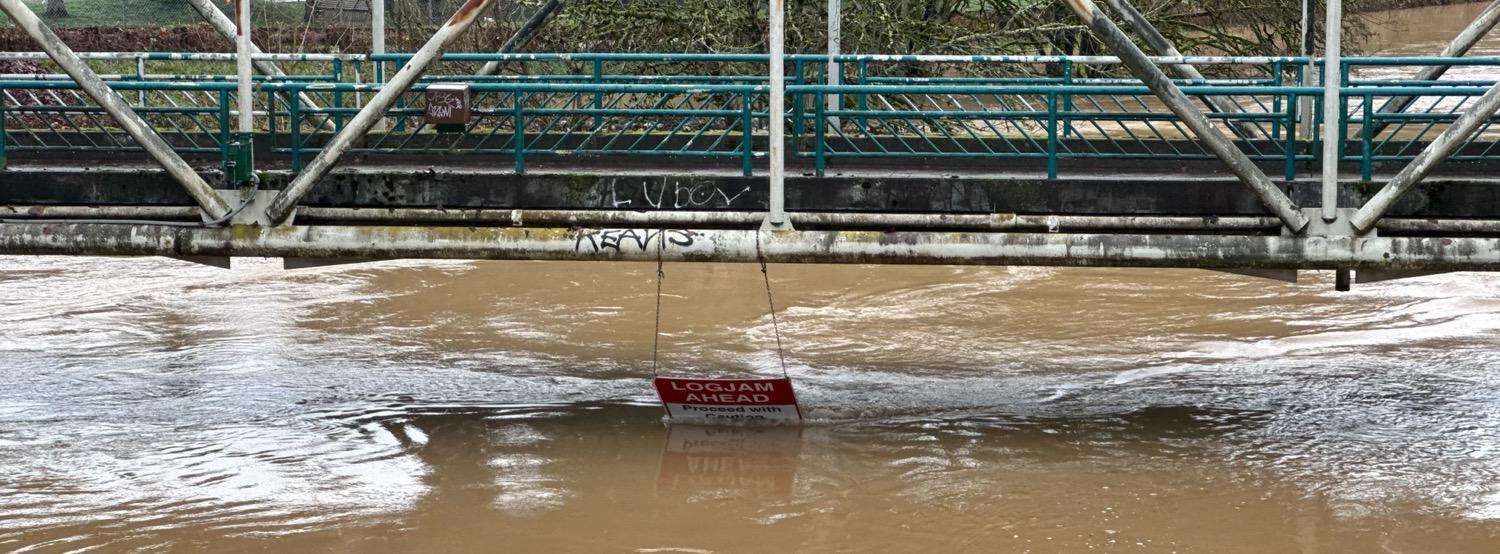 IMG_5465.jpeg A pedestrian bridge over a river with a sign that reads “Logjam Ahead, Proceed With Caution” hanging under the bridge and partially submerged in the flowing water.