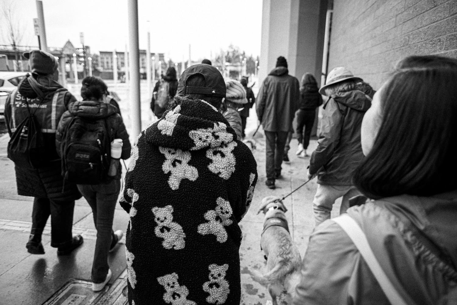 Light Rail Federal Way Extention Accessibility Tour - 002.jpg Black and white shot of a group of people walking along a sidewalk as seen from behind. Several have guide dogs, one has a white cane for the blind and is walking arm-in-arm with another person, and the person centered in the frame is wearing a fuzzy coat with a teddy bear pattern.