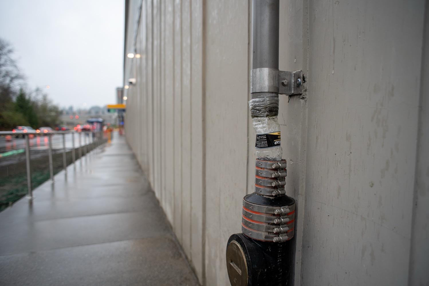 Light Rail Federal Way Extention Accessibility Tour - 037.jpg A concrete wall stretches into the distance, in the foreground is a PVC water drain pipe with a six-inch section missing that has a cut-up plastic water bottle being used as an improvisational fix.
