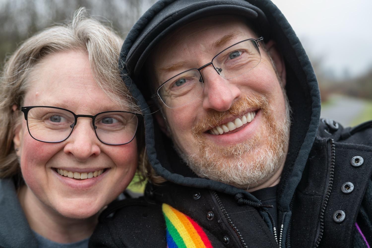 First Walk of 2026 - 005.jpg Selfie of me, a white man with greying red beard, weraing glasses and a black coat and hoodie with the hood up, and my wife a white woman with wavy blonde hair and glasses, both of us smiling.