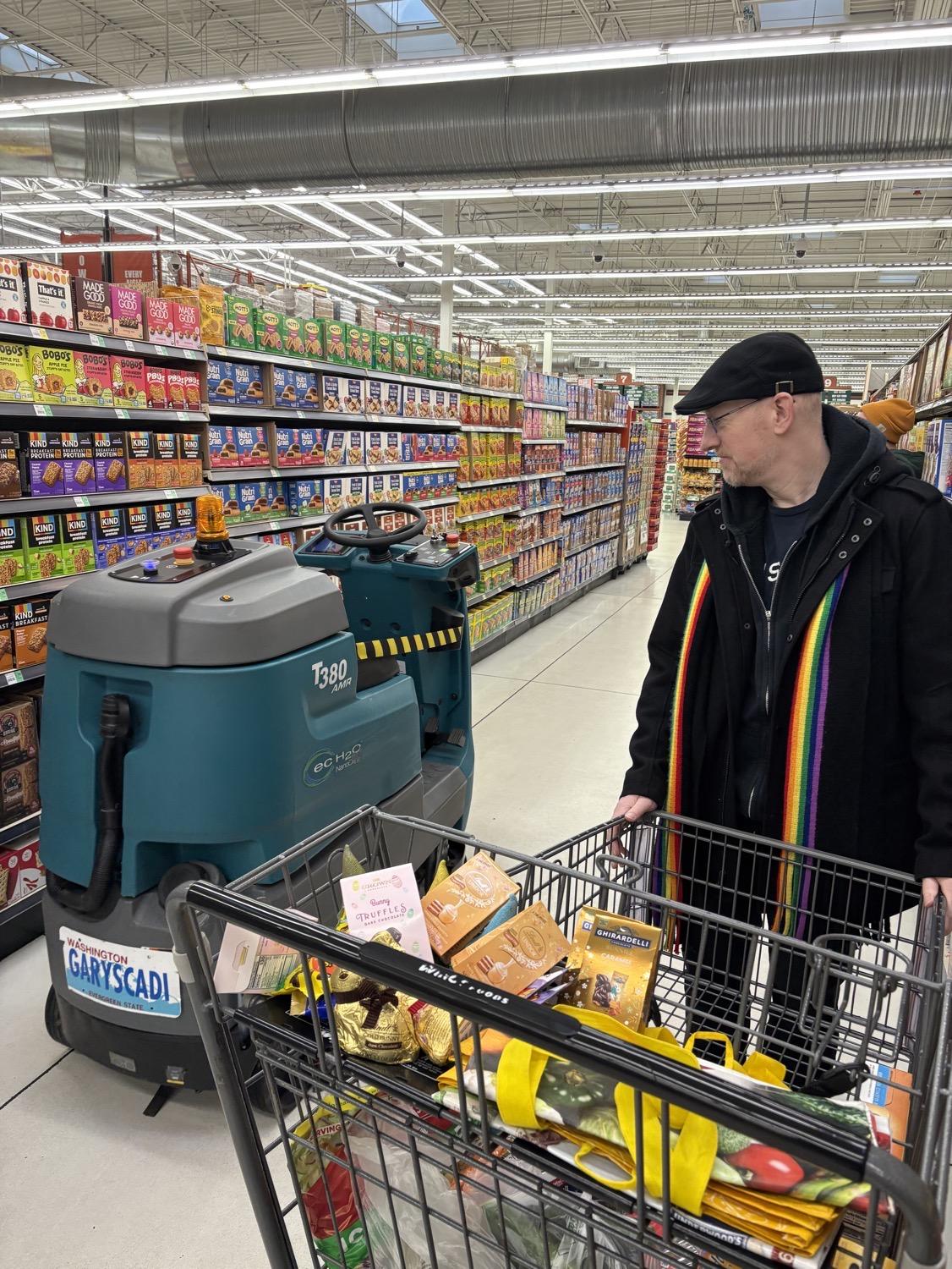 In a grocery store, I watch suspiciously as a robotic floor cleaner goes by our cart.