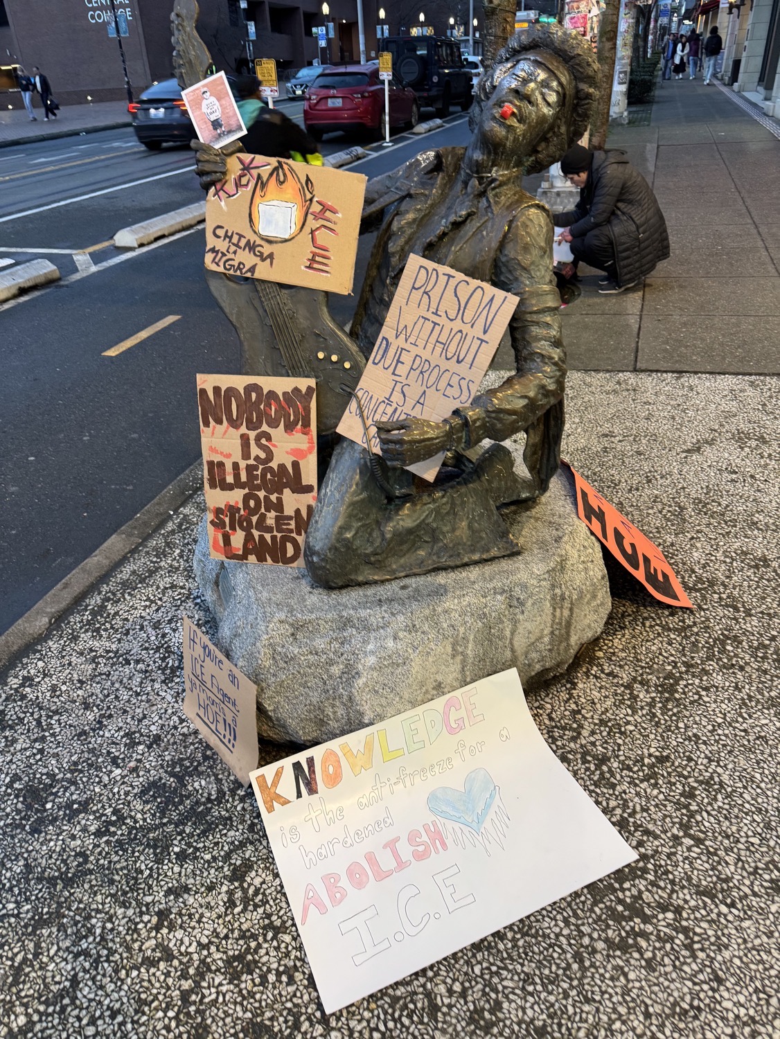A bronze stature of Jimi Hendrix on a city street, decorated with several protest signs and with a whistle placed in his mouth.
