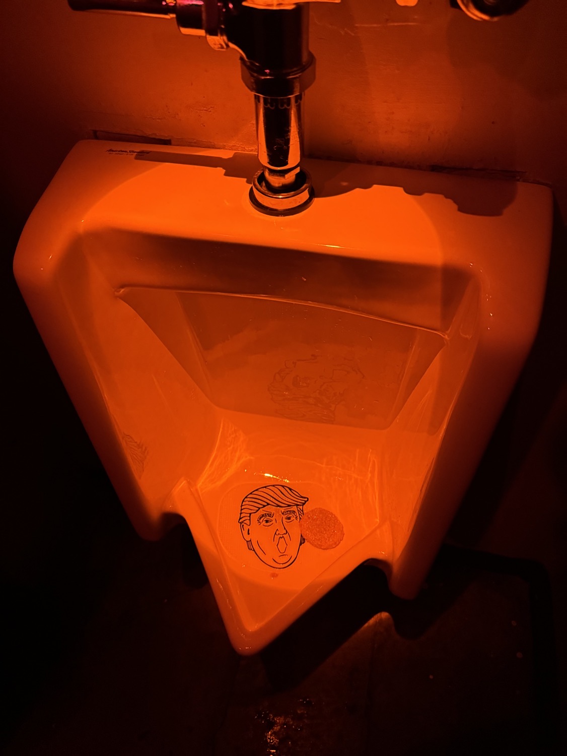 A urinal lit all in red, with an anti-splash mat featuring a drawing of Trump's face.
