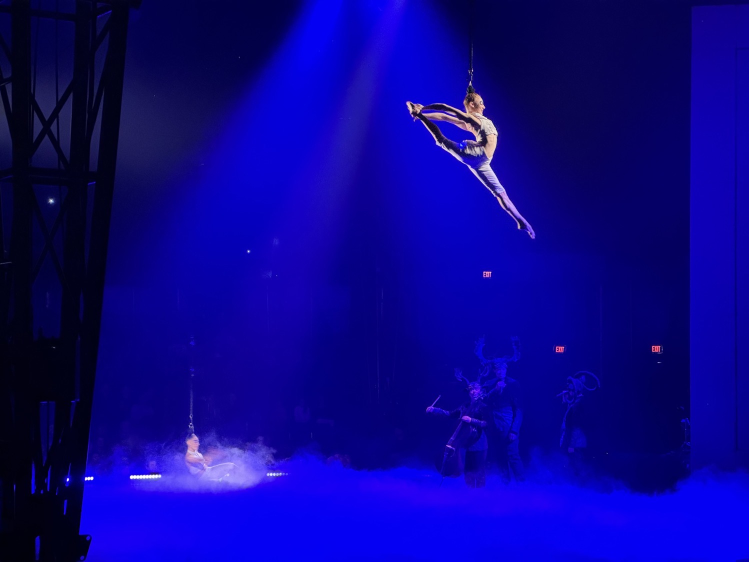 As musicians in dark outfits perform on a blue-lit stage, an acrobat hangs suspended in the air by her hair, legs stretched wide, one foot in front of her and her arms stretched back to hold her rear foot up behind her head.
