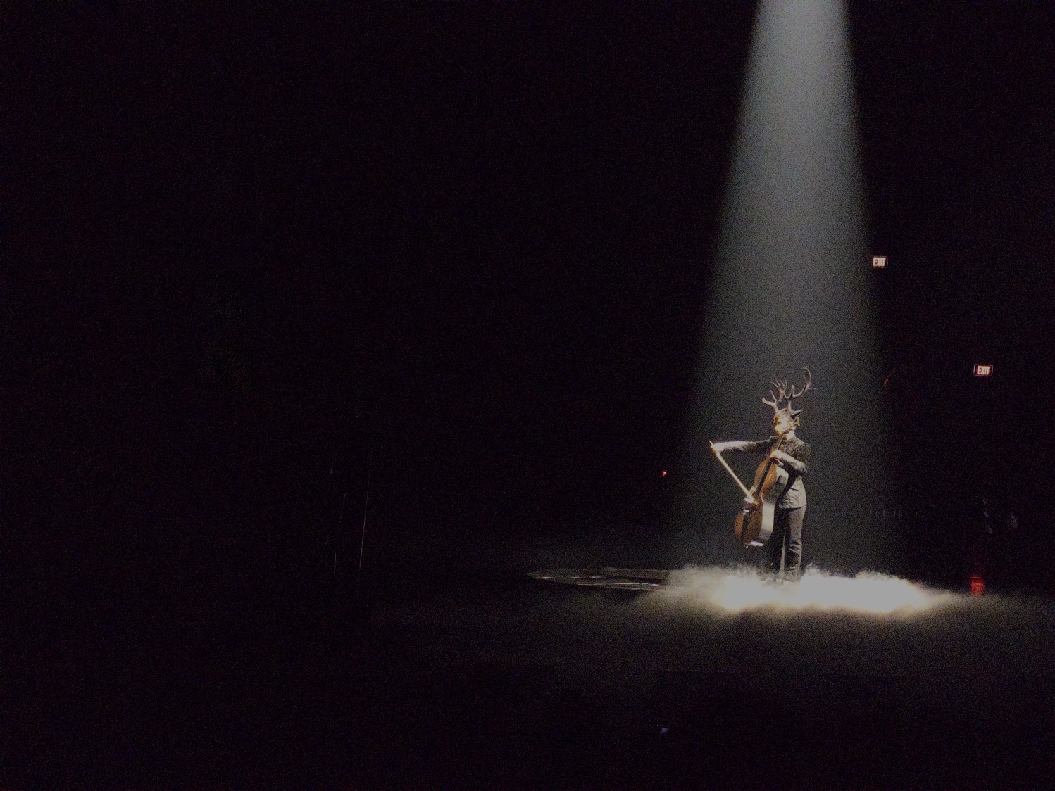 A musician stands in a spotlight on a dark stage, wearing antlers on their head and playing a cello.