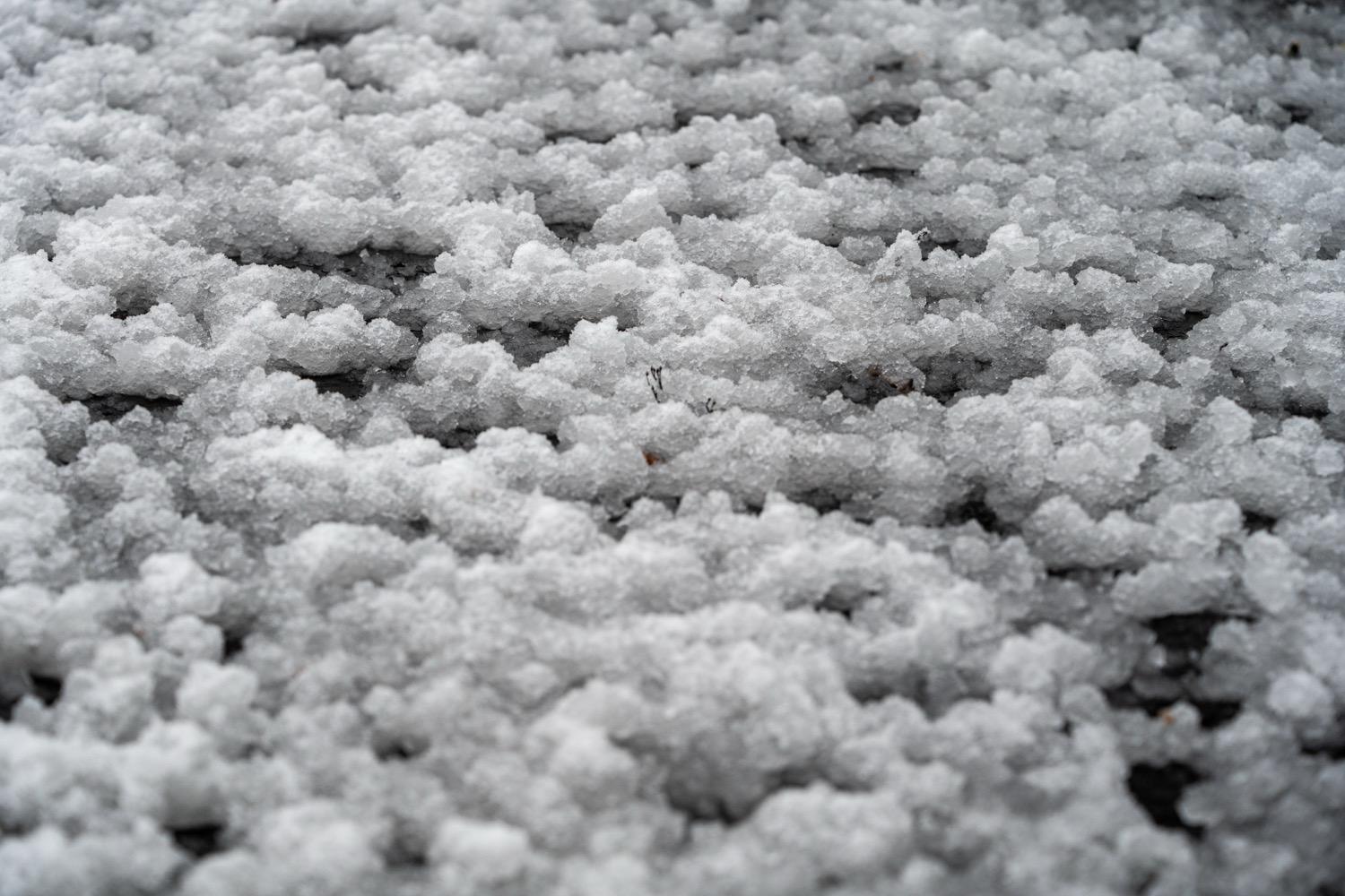 Pellets of slush and ice lie on the ground in a close-up, nearly monochrome shot, looking like a rough landscape.