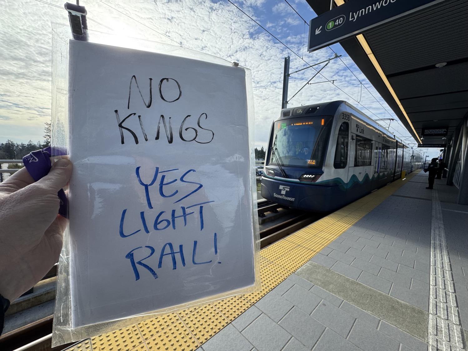 My hand holding up a sign that says 'no kings, yes light rail' as a light rail train pulls into the station.